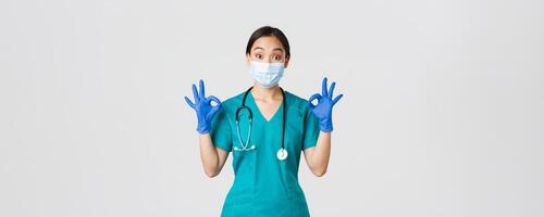 Covid-19, coronavirus disease, healthcare workers concept. Impressed asian female doctor, physician in medical mask, rubber globes and scrubs show okay gesture, compliment, praise excellent work photo