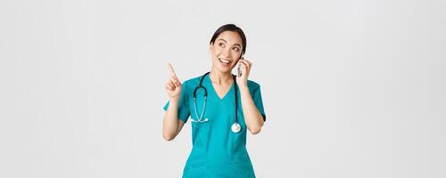 COVID-19, cuidado de la salud trabajadores y prevenir virus concepto. retrato de joven bonito asiático hembra doctor, enfermero en matorrales haciendo en línea orden, hablando en teléfono y señalando Superior izquierda esquina, sonriente foto