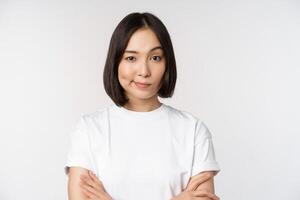 Close up of korean girl, looking skeptical, cross arms on chest and smirk, stare with disbelief at camera, standing over white background photo