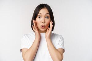 Close up portrait of asian girl looking surprised, wow face, reacting amazed at smth, standing in white tshirt over studio background, isolated photo