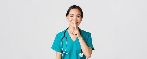 Healthcare workers, preventing virus, quarantine campaign concept. Smiling pretty asian doctor, nurse in scrubs smiling, shushing, asking stay quiet, standing white background photo