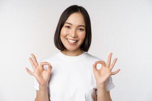 Very well, excellent. Smiling asian woman showing okay sign, approval, ok gesture, looking satisfied, recommending smth, standing over white background photo