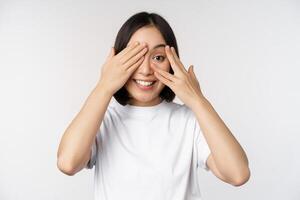 Portrait of asian woman covering eyes, waiting for surprise blindfolded, smiling and peeking at camera, standing over white background photo