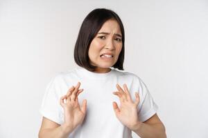 Disgusted asian woman rejecting smth, grimacing from dislike and aversion, stare with cringe, refusing offer, standing over white background photo