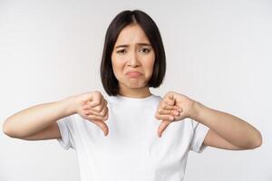 Image of asian woman showing thumbs down, dislike smth, looking disappointed, standing over white background photo