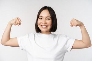 Smiling asian woman showing flexing biceps, muscles strong arms gesture, standing in white tshirt over white background photo