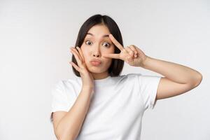 Cute asian girl posing with kawaii v-sign, peace gesture near face, standing in tshirt over white background photo