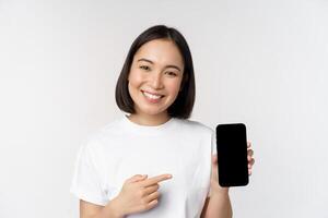 Smiling asian woman pointing finger at smartphone screen, showing application interface, mobile phone website, standing over white background photo