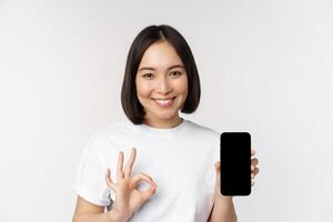 Smiling asian woman showing okay, mobile phone screen, recommending smartphone app, standing over white background photo