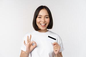 sonriente coreano niña demostración bueno firmar y recomendando crédito tarjeta de Copiar espacio banco, en pie en camiseta terminado blanco antecedentes foto