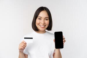 Portrait of smiling young asian woman showing mobile phone screen and credit card, standing over white background photo