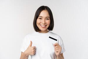 Portrait of beautiful young modern asian woman, showing credit card and thumbs up, recommending contactless payment, standing over white background photo