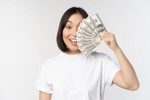 Portrait of smiling asian woman holding dollars money, concept of microcredit, finance and cash, standing over white background photo