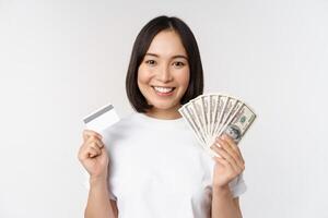 retrato de asiático mujer sonriente, participación crédito tarjeta y dinero dinero en efectivo, dólares, en pie en camiseta terminado blanco antecedentes foto