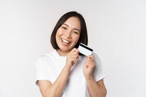 Image of smiling asian woman hugging credit card, buying contactless, standing in white tshirt over white background photo