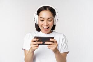 Happy asian woman in headphones, looking at smartphone, watching video on mobile phone and smiling, standing over white background photo