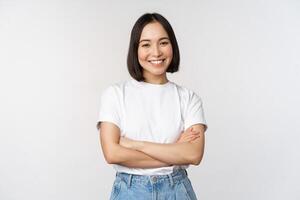 retrato de contento asiático mujer sonriente, posando seguro, cruzar brazos en pecho, en pie en contra estudio antecedentes foto