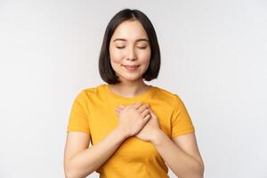 Romantic asian girfriend, holding hands on heart, smiling with care and tenderness, standing in yellow tshirt over white background photo