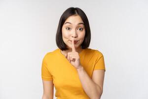 Keep quiet. Cute asian woman make shhh gest, showing shush, hush sign, press finger to lips, silence, standing over white background photo