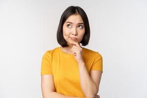 Image of thinking asian woman, looking aside and pondering, making decision, standing in yellow tshirt over white background photo