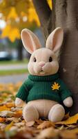 AI generated Photo Of A Stuffed Animal Bunny In A Sweater Leaning Against A Tree In A Est With Leaves On The Ground And A Yellow Maple Leaf In The Eground. AI Generated