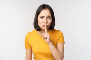 Portrait of angry asian woman shushing, press finger to lips, taboo stop talking sig, looking annoyed, scolding, standing over white background photo