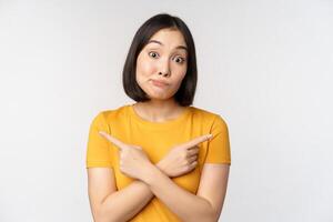 Complicated asian girl pointing fingers sideways, showing left and right choices, staring indecisive clueless, standing over white background photo