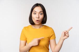 Amazed young asian woman, showing advertisement aside, pointing fingers right at promotion text, brand logo, standing happy against white background photo