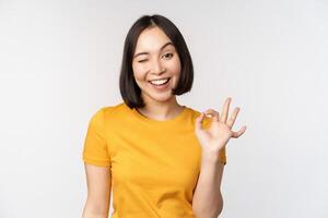 Beautiful young woman showing okay sign, smiling pleased, recommending smth, approve, like product, standing in yellow tshirt over white background photo