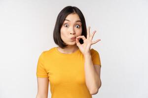 Cute asian girl seal lips, zipping mouth with finger, promise to keep secret, taboo gesture, standing in yellow tshirt over white background photo