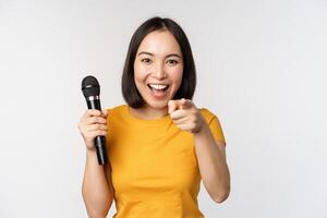 entusiasta asiático niña con micrófono, señalando dedo a cámara, sugerencia usted a cantar, en pie terminado blanco antecedentes foto