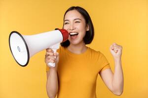 Announcement. Happy asian woman shouting loud at megaphone, recruiting, protesting with speaker in hands, standing over yellow background photo