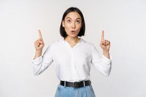 Enthusiastic asian business woman pointing, looking up with happy smiling face, showing company logo or banner, standing over white background photo