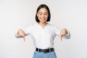 Portrait of happy asian woman pointing fingers down and looking below at advertisement, showing info banner, advertising, standing over white background photo