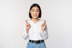 Portrait of young korean woman, asian girl cross fingers and praying, making wish, anticipating, waiting for results, standing over white background photo