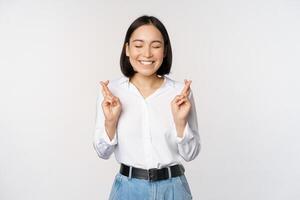 Portrait of young korean woman, asian girl cross fingers and praying, making wish, anticipating, waiting for results, standing over white background photo