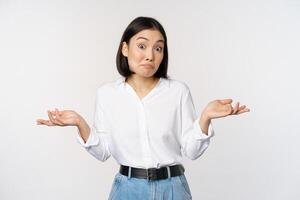 Image of confused asian office manager, office lady shrugging shoulders and looking clueless, standing puzzled against white background photo