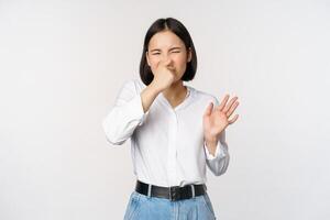 Disgusted asian girl close nose and showing rejection gesture, digusting bad smell, standing over white background photo