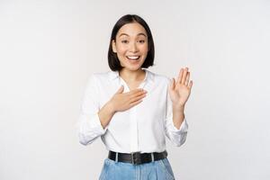 Image of cute young female office worker, asian girl student raising hand up and put palm on chest, name herself, introduce, making promise, standing over white background photo