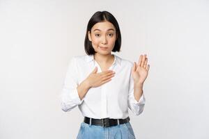 Image of cute young female office worker, asian girl student raising hand up and put palm on chest, name herself, introduce, making promise, standing over white background photo