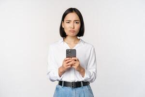 Asian woman holding smartphone and looking with doubt, disappointed with mobile phone app, standing agaist white background photo