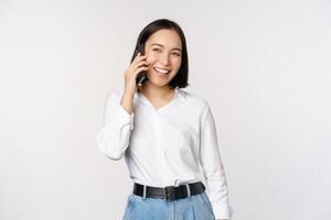 simpático sonriente asiático mujer hablando en teléfono, niña en llamar, participación teléfono inteligente y reír, discurso, en pie terminado blanco antecedentes foto