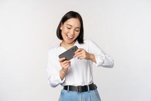 Young korean woman, asian girl playing mobile video game on smartphone, looking at horizontal phone screen, standing over white background photo