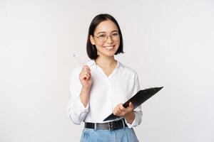sonriente joven asiático mujer tomando notas con bolígrafo en portapapeles, mirando feliz, en pie en contra blanco antecedentes foto