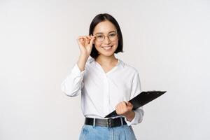 joven mujer, oficina trabajador gerente en anteojos, participación portapapeles y mirando me gusta profesional, en pie en contra blanco antecedentes foto