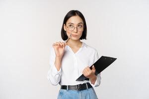 Asian girl in glasses thinks, holds pen and clipboard, writing down, making notes, standing over white background photo