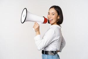 Beautiful young asian woman talking in megaphone, screams in speakerphone and smiling, making announcement, shout out information, standing over white background photo