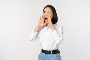 Image of young asian woman calling for someone, shouting loud and searching around, standing against white background photo