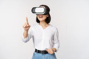 Amazed young asian woman using vr headset. Korean girl in virtual reality glasses pointing at smth and smiling, white background photo