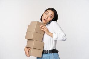 Young asian businesswoman answer phone call, talking on mobile while carrying pile of boxes with orders, standing over white background photo